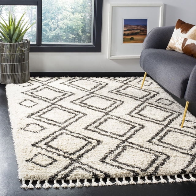 Safavieh Moroccan Fringe Kaelea Geometric Shag Area Rug with plush white synthetic yarns and black diamond geometric pattern, featuring fringed edges, displayed in a modern room with a gray armchair, cowhide pillow, potted plant, and large window, ideal for accent or casual decor.