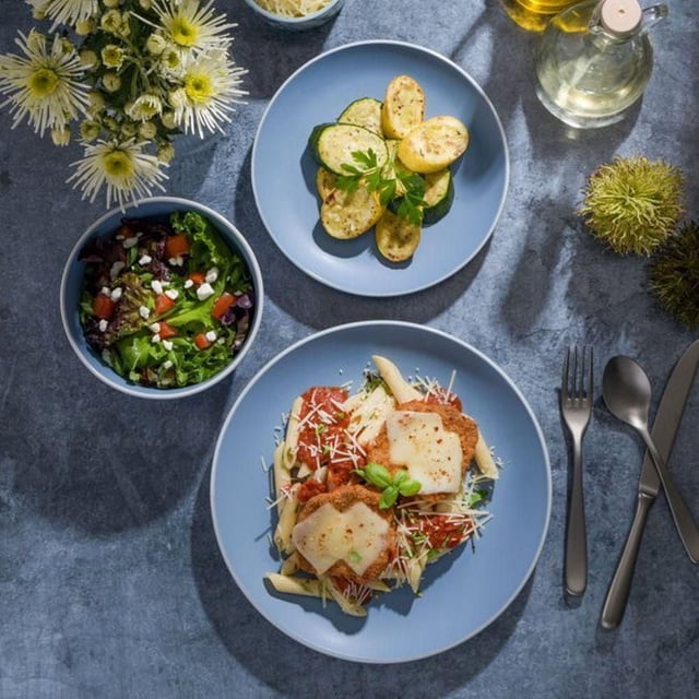 Tia Mowry 12 Piece Stoneware Dinnerware Set, Blue with a top-down view of a blue matte finish dinner plate holding penne pasta topped with breaded chicken and melted cheese, a smaller blue plate with grilled zucchini and potatoes garnished with parsley, and a blue bowl of mixed salad, arranged on a grey textured surface with stainless steel cutlery, a glass bottle of oil, and white flowers nearby.