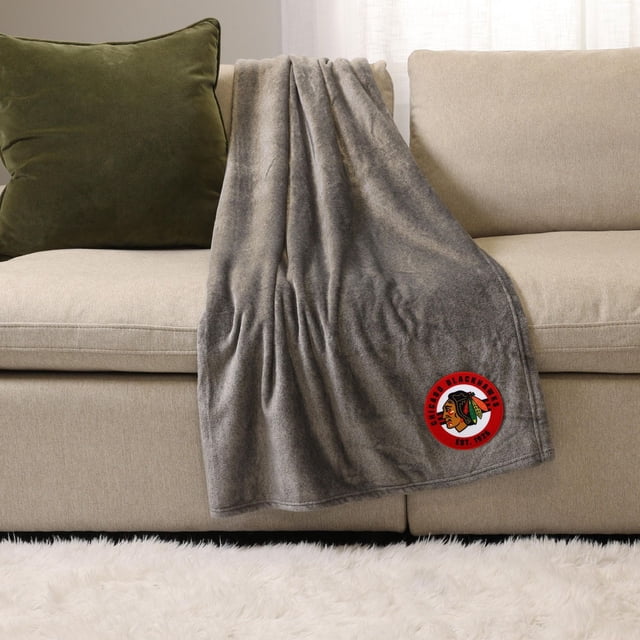 NHL, NBA, MLB Grey Sports Throw Blanket, 50" x 60", draped over a beige sofa with a green pillow, made of soft plush 100% polyester featuring a red Chicago Blackhawks logo patch, ideal for indoor warmth and cozy lounging.