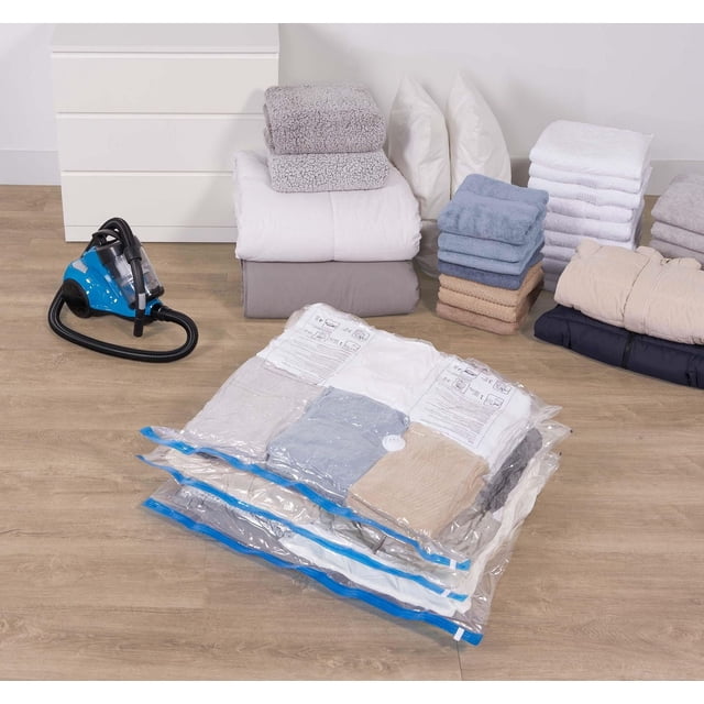 Mainstays 3 piece Extra Large Cube Storage Bags vacuum-sealed with bedding and towels inside on a wooden floor near a blue vacuum cleaner and folded blankets, pillows, and towels, providing a space-saving storage solution that protects items from dust, moisture, and odors, also suitable for storing seasonal clothing.