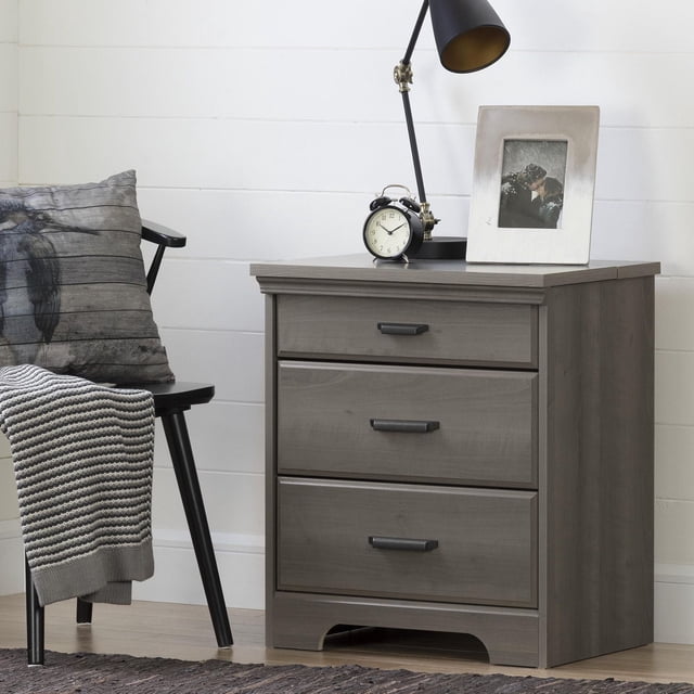 South Shore Versa Nightstand with Charging Station and Drawers for Adult in gray maple, front view showing three drawers with black metal handles and a sliding top charging station for device cord management, styled with a black adjustable desk lamp, vintage alarm clock, and photo frame, positioned next to a black chair with a gray pillow and knitted throw, set against white shiplap walls and light hardwood flooring.