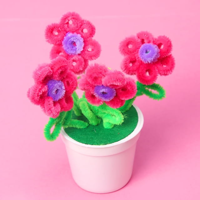 Vaidha DIY Hairy Flower Potted Craft Kit for Kids featuring a mini plastic flower pot with colorful hair roots shaped into vibrant pink and purple flowers with green stems. The craft kit is designed for creative activities like Mother's Day twisted stick projects. The pot measures approximately 14cm in height and 3.5cm in diameter. Displayed against a pink background.