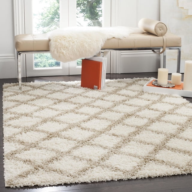 Safavieh Dallas Jerrie Tapis Shag Géométrique pour Aire avec un Motif de Losanges Pelucheux en Crème et beige, Posé sur un Plancher de Bois Franc Foncé sous un Banc en Cuir beige avec des Pattes Chromées, Accentué d'Un Jeté en Fausse Fourrure Blanc et d'Un Coussin Cylindrique, avec des Livres et des Bougies à Proximité, Convient pour Améliorer la texture et la Chaleur dans les Intérieurs Modernes.