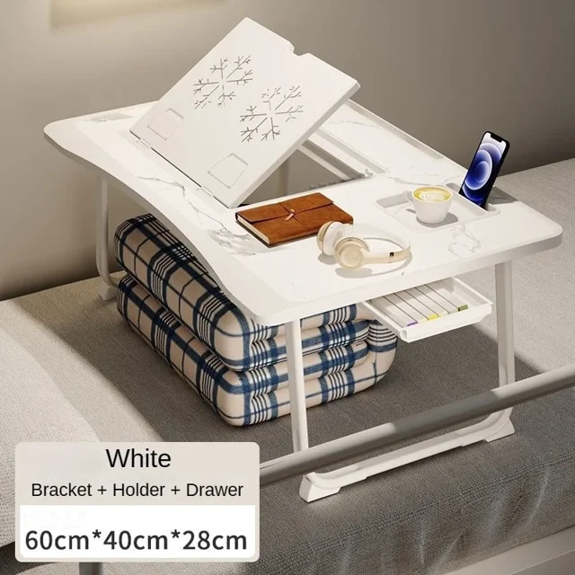 Bureau d'Ordinateur Portable Pliable pour Lit avec Support à Hauteur Réglable à 6 Niveaux, Illustré en Fini Blanc avec un Motif Flocon de Neige sur la surface. le Bureau Rectangulaire Mesure 60x40x28 cm et Comprend un Tiroir Intégré, un Support pour Téléphone et un Porte-Gobelet. Il Est Posé sur un Lit avec des Coussins à Carreaux en Dessous, Mettant en Valeur Son Utilisation comme table de Chevet pour Travailler, Jouer Ou Manger. le Bureau Est Pliable et Conçu pour un Rangement compact.