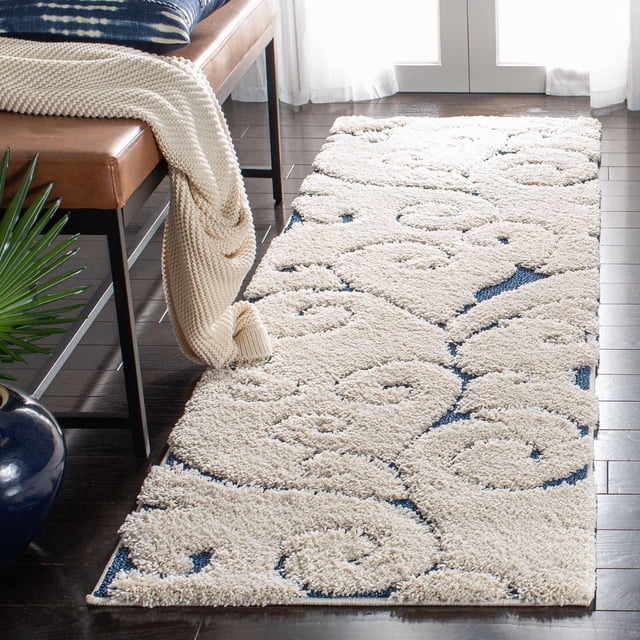 SAFAVIEH Florida Douglas Floral Vines Shag Area Rug runner with crème tone-on-tone sculpted high-low pile featuring damask vine patterns over blue base, placed on dark hardwood floor beside a leather bench with cream knit throw and blue cushion near bright window, ideal for adding softness and texture to narrow spaces.