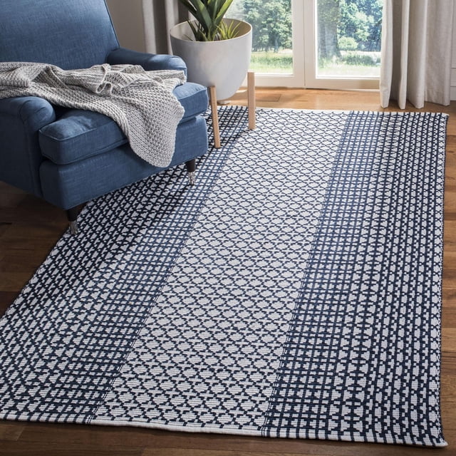 Safavieh Montauk Cara Geometric Area Rug featuring flat-weave cotton with navy and white geometric patterns in vertical stripes, placed on a hardwood floor beside a blue armchair with a knitted throw, near a window with beige curtains and a potted plant. Ideal for coastal and contemporary home décor.