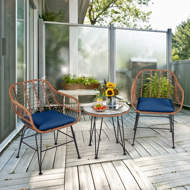 Costway 3PCS Patio Rattan Bistro Set with two brown rattan armchairs featuring navy cushions and a round tempered glass coffee table. The set is arranged on a wooden patio with plants and sunflowers in the background, suitable for outdoor areas like patios, gardens, or balconies.