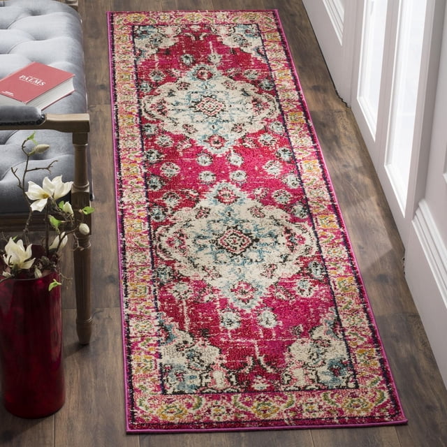 Safavieh Monaco Toria Traditional Area Rug with vibrant pink, cream, and blue Persian-inspired patterns, power-loomed polypropylene with mixed high and low loop pile, shown on a dark wood floor beside a gray tufted bench and red vase with white flowers, ideal for adding Bohemian-chic style to hallways or entryways.