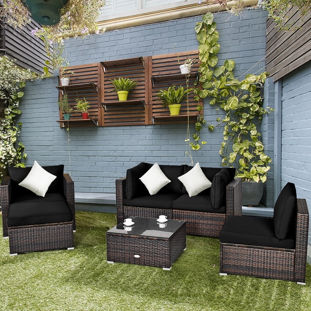 Costway 6PCS Patio Rattan Furniture Set Cushion Sofa Coffee Table Ottoman in black, featuring 2 corner sofas, 1 armless sofa, 1 single sofa, and 2 ottomans. Made with durable PE wicker and powder-coated steel for outdoor use. Includes thick, washable cushions and a tempered glass coffee table. Shown in a backyard setting with green grass and a blue brick wall adorned with plants.