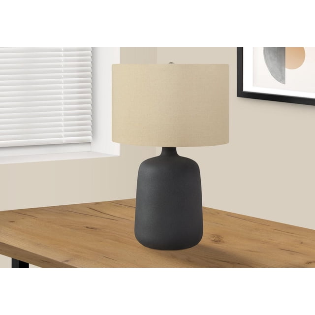 Monarch Specialties Lighting 24-inch table lamp with a black ceramic textured vase-shaped base, beige linen drum shade, and silver finial, featuring a 3-way rotary switch for adjustable lighting, placed on a wooden table near a window with white blinds and abstract wall art in a neutral room.