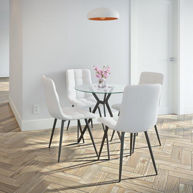 CorLiving Eliana Contemporary 5-Piece Compact Round Glass Indoor Dining Table Set with 4 white velvet-upholstered chairs featuring tufted backs and black iron legs, placed on light wood herringbone flooring beneath a white pendant light, with a clear tempered glass tabletop holding a small vase of pink flowers, suitable for compact dining areas or breakfast nooks.