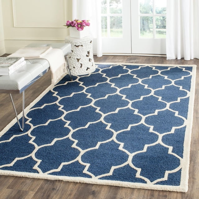 Safavieh Cambridge Kevin Geometric Area Rug in blue with white geometric trellis pattern, hand-tufted wool with plush cut pile and texture-rich high/low loop and cut piles, on wood floor in a bright room with white curtains, a white decorative side table with pink flowers, and a gray bench with books and a beige throw. Often used to add modern American style and warmth to home or office decor.