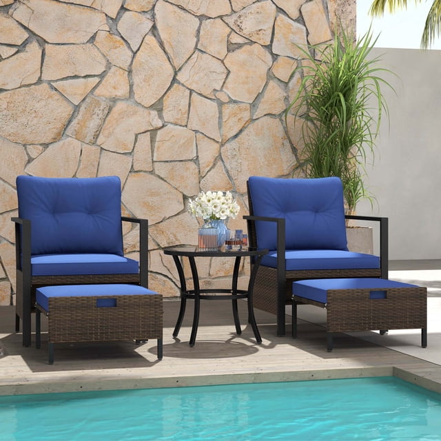 Costway 5 PCS Outdoor Furniture Set with two mix brown PE rattan armchairs featuring navy cushions, two versatile ottomans, and a round tempered glass coffee table. Ideal for patio conversation sets, this arrangement is displayed poolside against a stone wall, highlighting its stylish and durable design.