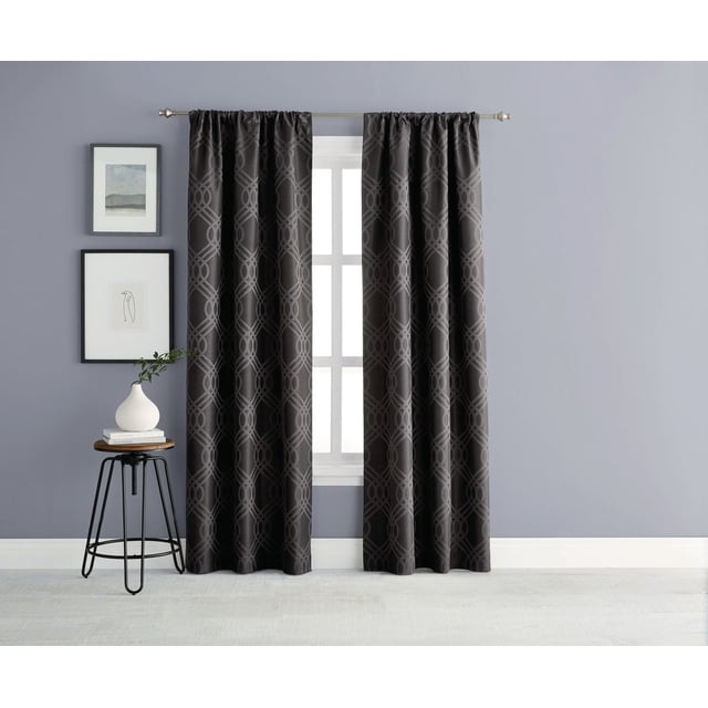 Eclipse Zana Trellis 63" Blackout Rod Pocket Window Panel in black with a wavy interlocking geometric jacquard pattern, hanging on a silver rod over a white window frame. The blackout curtain is 100% polyester with premium thermal lining for light blocking, privacy, and energy savings. Rod pocket style drapes to the floor in a room with grey walls, a small round wooden side table with black metal legs, a white vase with greenery, and two framed minimalist artworks.