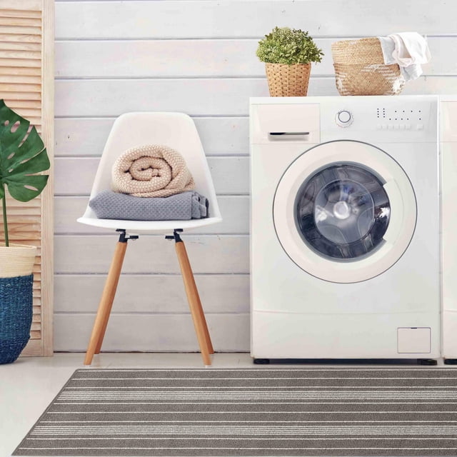2 ft x 5 ft Olin Grey Mat with horizontal light grey and white stripes placed in front of a white front-load washing machine in a laundry room. The machine-tufted polypropylene mat protects floors from dirt, salt, and stains. A white chair with wooden legs holds folded beige and grey towels, with a woven basket and green plant on the washing machine.