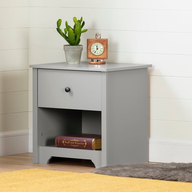South Shore Vito 1-Drawer Nightstand in soft gray with a single drawer featuring a black metal knob and an open lower shelf holding a book titled 'Stuff and Nonsense', topped with a potted green cactus and a wooden vintage clock, positioned on a light wood floor against a white paneled wall with a yellow rug and gray fur accent, suitable for bedside storage and organization.