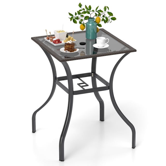 Gymax Table de Bar Bistro Carrée de 28,5 Po avec Trou pour Parasol de 2 Po et Dessus de Table en Verre Sérigraphié. la Table Présente un Cadre en Rotin PE Brun Mélangé et une surface en Verre Résistante à la Chaleur et aux Rayures. Elle Est Supportée par un Cadre en Métal Enduit de Poudre avec Raccords Vissés Renforcés et Patins de Pieds Ajustables pour la Stabilité. la Table Est Présentée sur un Fond Blanc avec des Objets Décoratifs comme un vase, des Collations et une Tasse à Café Posée sur le Dessus de Table.