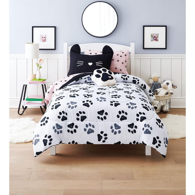 Mainstays Kids MEOW Bed in a Bag twin bedding set with a white comforter featuring black and gray paw prints, a black cat face pillow with ears, a pink pillow with small paw prints, and a white paw-shaped decorative pillow, displayed in a child's room with light hardwood floors, white wainscoting, a round mirror, framed cat artwork, a side table with a lamp and books, and plush toys in a basket, suitable for adding playful style and comfort to kids' bedrooms.