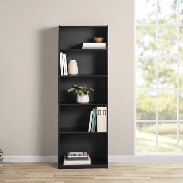 Mainstays 5-Shelf Bookcase with Adjustable Shelves in black wood grain finish, featuring a closed-back design with three adjustable shelves and two fixed shelves, holding books, a white ceramic pot with a green plant, a beige vase, and office binders, positioned against a beige wall near a window with natural daylight, suitable for home office or living room storage and display.