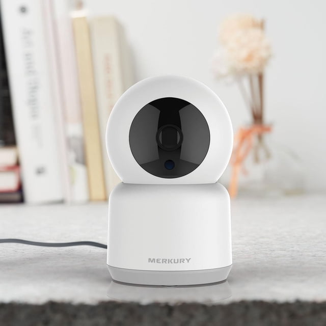 Merkury Innovations Smart Wi-Fi Auto Follow Camera HD 1080P with a white rounded base and black circular lens, front-facing on a light surface with the Merkury logo visible, set against a blurred background of books and a flower vase, ideal for indoor smart home security with 1080p live streaming, motion alerts, and auto-following features.