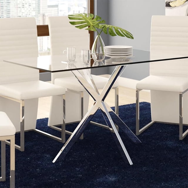 Home Gear Dining Table with a 31.5 by 51.25 inch clear tempered glass tabletop featuring rounded edges, supported by a chrome-finished X-frame steel base with flat square platforms and felt foot pads. The table is on a dark blue rug surrounded by white modern dining chairs with chrome legs, with a glass vase holding green leaves and stacked white plates on top, ideal for intimate dinners or family meals.