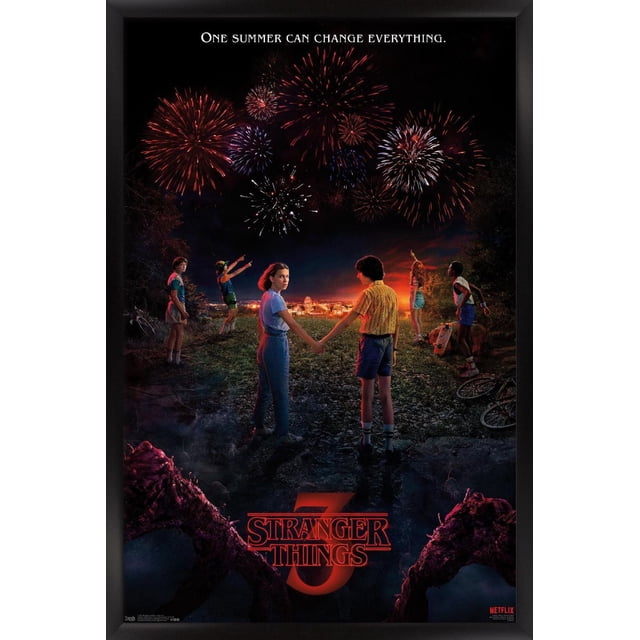 Netflix Stranger Things: Season 3 - Key Art Wall Poster depicting six children standing on a rocky path at night watching fireworks over a brightly lit town. Two children hold hands center frame while others point at the sky. The poster features the tagline 'One summer can change everything' at the top and the red Stranger Things 3 logo at the bottom, framed in black.