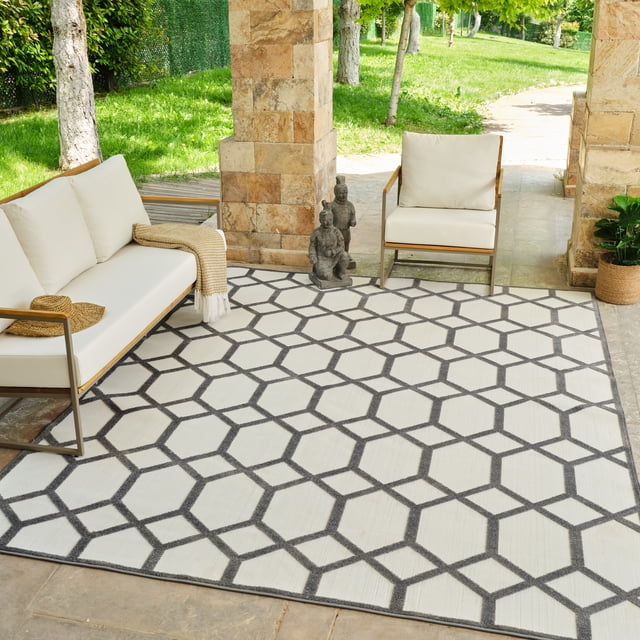 Rug Branch Alfombra Abstract Indoor Outdoor Area Rug, Grey Beige, Geometric - A front-facing view of a grey and beige geometric patterned area rug with interconnected hexagonal shapes on a stone patio. The rug is complemented by white cushioned outdoor seating with wooden armrests, positioned between stone pillars with a green lawn and trees visible beyond. Ideal for high-traffic outdoor areas, made from durable polypropylene with jute backing for strength.