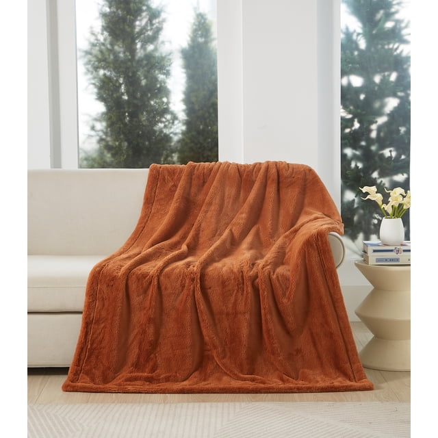 Better Homes & Gardens Faux Fur Throw in copper, oversized 50 x 72 inches, draped over a beige sofa in a bright room with large windows showing evergreen trees. The reversible throw features plush faux fur on both sides and is placed beside a beige side table with a white vase of flowers and stacked books. Ideal for cozying up on the couch or adding warmth to a bed.