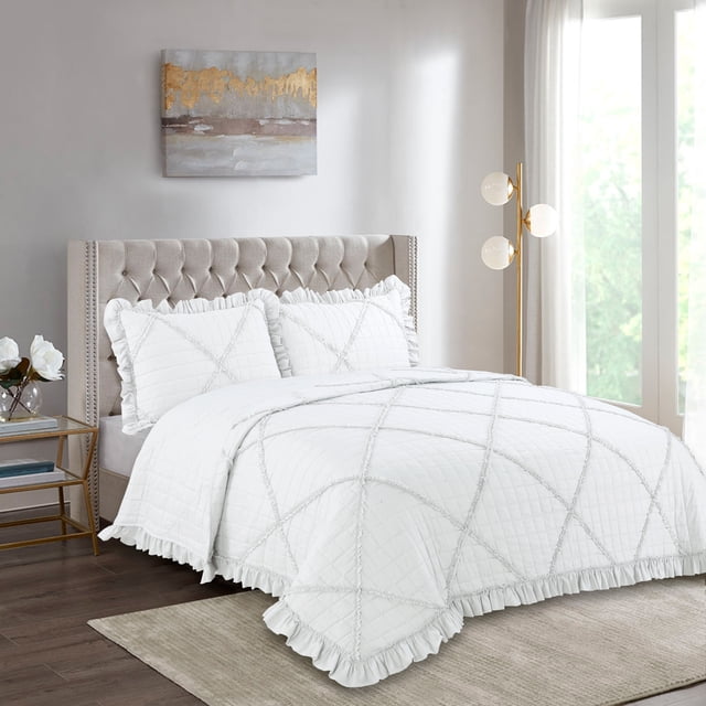 Marina Decoration Lush Ruffled Textured Shabby Chic Stitching Coverlet Bedspread Ultra Soft Solid Summer Embroidered Quilt Set with Quilted Shams in white on a beige tufted bed. The breathable quilt features embroidered large square ruffle patterns and matching ruffled pillow shams. Set in a sunlit bedroom with a gold floor lamp, glass side table with white flowers, and a neutral rug on dark hardwood floors. Suitable for all-season use and often used as a lightweight blanket.