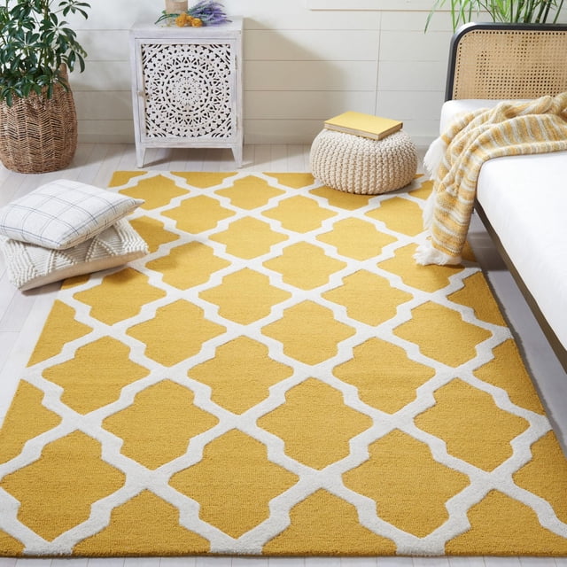 Safavieh Cambridge Liam Geometric Area Rug in yellow and white with a repeating trellis pattern, hand-tufted wool texture, placed on light wood flooring in a bright room with a white carved cabinet, woven pouf with a yellow book, and a sofa with a yellow plaid throw blanket, suitable as an accent rug for home decor.