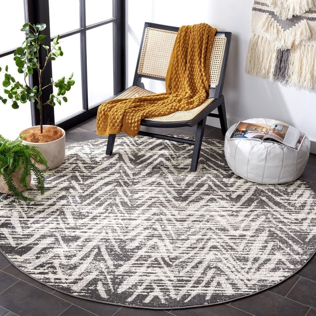 SAFAVIEH Cottage Tapis d'Intérieur/extérieur à Chevrons Ronni Gris avec Motif à Chevrons, Posé sur un Plancher de Tuiles Foncées dans une Pièce Lumineuse. le Tapis Rond Est Fait de Fibres Synthétiques Durables Idéal pour une Utilisation Intérieure et Extérieure. une Chaise en Bois Noire avec Siège et Dossier en Canne Tressée Tient un Jeté Tricoté Jaune Moutarde. un pouf en Cuir Blanc avec un Magasin Ouvert s'Asseoit à Proximité, Accompagné de Deux Plantes en Pot Ajoutant des accents Naturels.