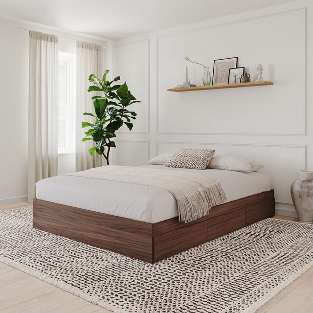 Nexera Alibi Lit de Rangement à 3 Tiroirs en Fini Stratifié Noyer avec Trois Tiroirs de Rangement Réversibles sur Glissières en Métal, Montré dans une Chambre Minimaliste avec des Murs Blancs et de Légers Planchers de Bois Franc. le Cadre de Lit Supporte un Matelas standard Sans Sommier et Est Stylé avec une Literie Neutre, un Jeté Texturé et un Coussin à Motifs. la Chambre Présente une Grande Plante Verte Près d'Une Fenêtre avec des Rideaux Crème, une Tablette Flottante en Bois avec des vases Décoratifs et des Oeuvres Encadrées, et un Tapis à Motifs sous le Lit
