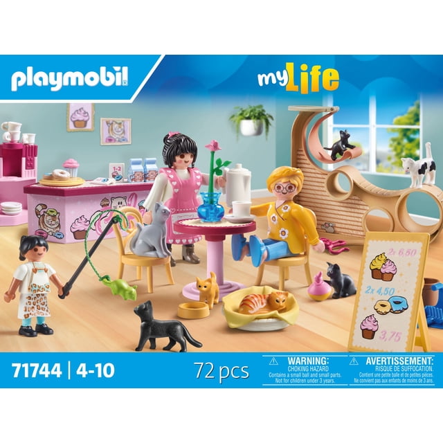 Playmobil Cat Cafe Play Set depicting a cafe scene with two figures seated at a pink round table with coffee and a flower vase, multiple cats playing and resting on chairs, floor, and a wooden cat playground with ramps and tunnels, a counter displaying cupcakes and donuts, and a menu board with prices, all arranged on a light wood floor with a window and hanging plant in the background.