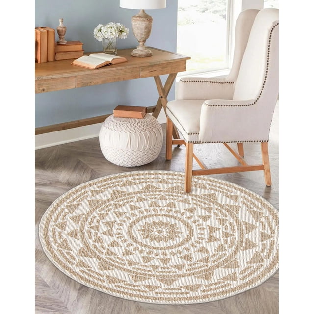 ECARPET Jute Style Area Rug for Bedroom, Living Room, Dining Room Finley Collection with a round jute style rug featuring a geometric pattern in sand and ivory tones, placed on light hardwood flooring beside a beige upholstered armchair with wooden legs and nailhead trim, a wooden desk with books and a lamp, and a white knitted pouf with a book on top, suitable for adding warmth and texture to various indoor spaces.