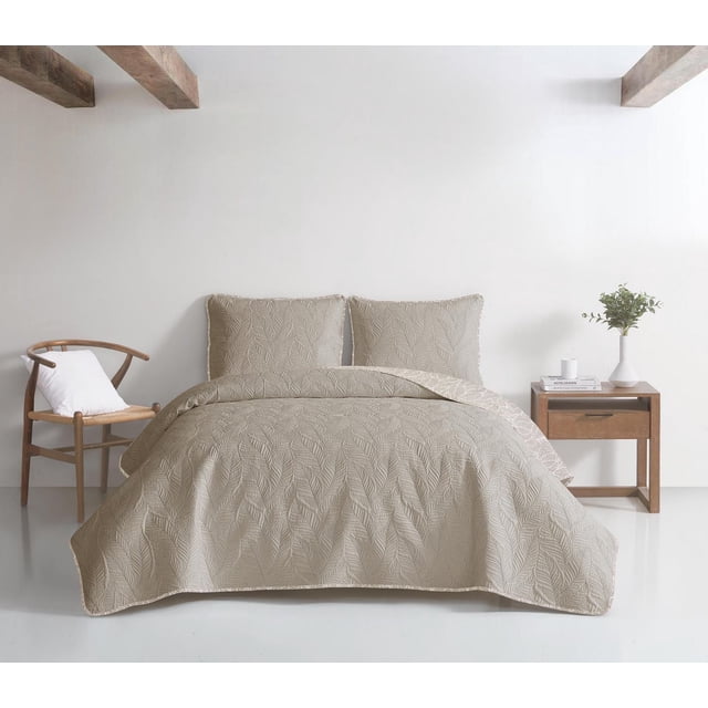 Delia Ensemble de Courtepointe Réversible 3 Pcs sur un Lit avec une Courtepointe en Microfibre beige Présentant un Motif de Feuilles Tropicales Cousues et Deux Taies d'Oreiller Assorties. la Courtepointe Se Renverse en une Impression Botanique Blanche et beige avec une Bordure Blanc sur beige. le Cadre Comprend une Chaise en Bois avec un Coussin Blanc à Gauche et une Table de Nuit en Bois avec un vase Blanc Tenant de la Verdure et des Livres à Droite, contre un Mur Blanc avec des Poutres en Bois Exposées.