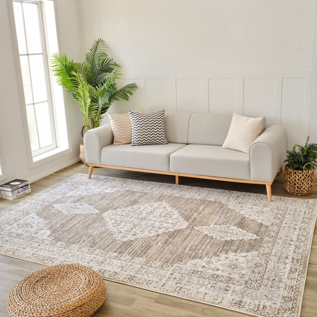 Rug Branch Nomad Vintage Indoor Area Rug in beige brown distressed pattern, 7'x10' (6'7" x 9'6"), displayed in a bright living room with a light grey sofa with wooden legs and three patterned cushions, a woven round ottoman, and green potted plants near large windows, ideal for living room, bedroom, dining room, or kitchen.