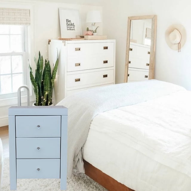 Nightstand with three drawers in light blue finish and clear round knobs, placed beside a bed with white bedding and gray throw in a bright bedroom with a white dresser, tall green plant, full-length mirror, and wall hat decor.