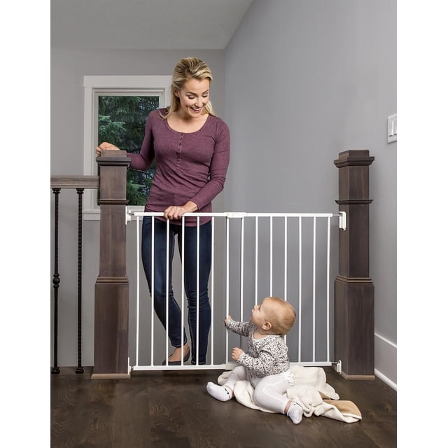 Regalo Barrière de Sécurité en Métal pour le Dessus des Escaliers en Métal Blanc Installée entre des Poteaux d'Escalier en Bois Foncé, Sécurisée avec Quincaillerie et Dotée d'Un Loquet Rotatif pour un Trajet Facile. une Femme Se Tient derrière la Barrière pendant Qu'un Bébé s'Asseoit sur une Couverture devant. la Barrière s'Adapte aux Ouvertures de 24 à 40,5 Pouces et Est Conçue pour Protéger les Enfants et les Animaux des Escaliers.