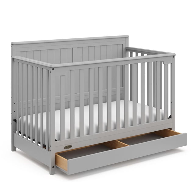 Graco Hadley 5-in-1 Convertible Baby Crib with Drawer in pebble gray, featuring a farmhouse-style design with vertical slats and paneled headboard, made from durable pine and engineered wood, with a full-size under-crib storage drawer divided into two compartments, shown with a white mattress.