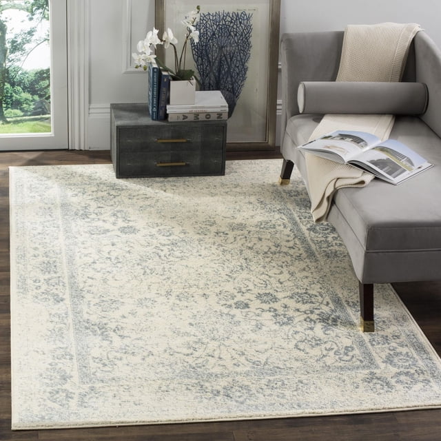 SAFAVIEH Adirondack Wyatt Traditional Area Rug with a rectangular shape and muted cream and light blue over-dye pattern on dark hardwood flooring. It complements a gray chaise lounge with brass-capped legs, beige throw, and an open book. A dark gray side table with brass handles holds books and a white orchid, with framed artwork behind it. Suitable for adding rustic lodge style to living spaces.