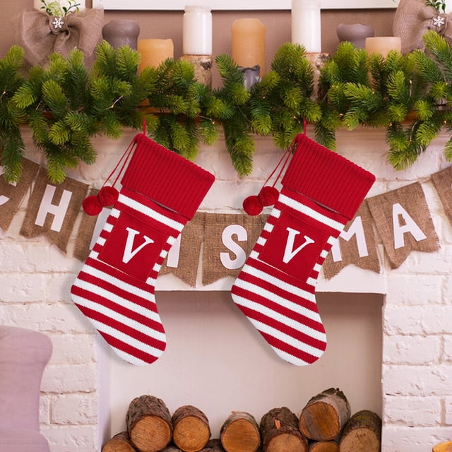 Christmas Large Letters Christmas Stockings by Czzoypi, featuring a festive red and white striped design with bold letter 'V' on the front. These polyester fiber stockings are hanging on a fireplace mantel decorated with greenery, candles, and a burlap Christmas banner. Perfect for holiday decor, they include a sturdy loop for easy hanging and spacious interiors for gifts and surprises.