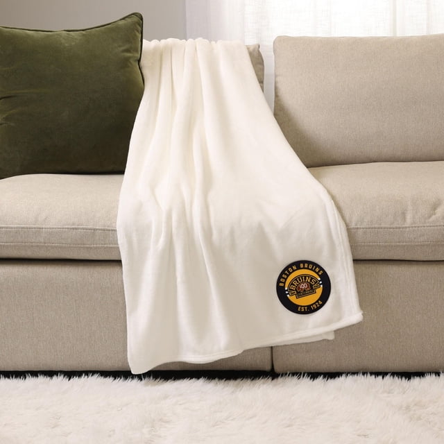 LNH , NBA, MLB Couverture de Sport Blanche, 50 Pouces x 60 Pouces, Peluche 100 % polyester, Drapée sur un sofa beige avec un Oreiller Vert Foncé, avec un Écusson logo des Boston Bruins en Noir et Jaune Près du Coin Inférieur, Idéale pour une Chaleur Intérieure Douillette et une Utilisation Polyvalente comme Jeté Superposé.