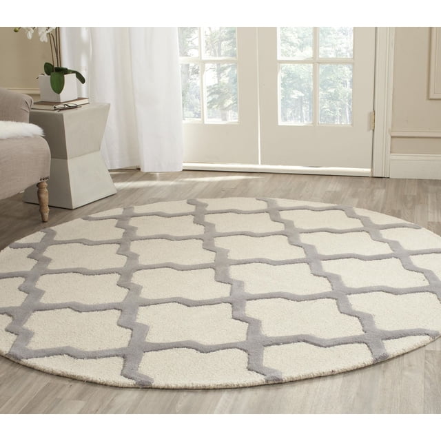 Safavieh Cambridge Liam Geometric Area Rug, round with cream base and light gray geometric trellis pattern, hand-tufted wool featuring plush cut pile and textured high/low loops, placed on light wood flooring in a bright living room with beige armchair and side table, ideal for adding warmth and classic style to home or office decor.
