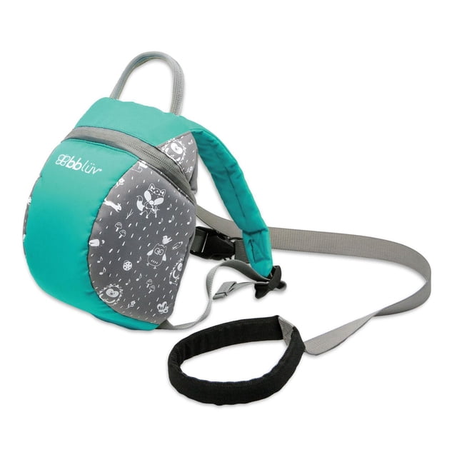 bblüv - Päk - Toddler Backpack (Aqua) shown in a 3/4 profile view with aqua and grey panels decorated with white animal and nature prints. Features padded shoulder straps, a top grab handle, and a removable safety rein with a padded black handle for parental control. Made of durable, washable fabric with an internal pocket and name tag inside.