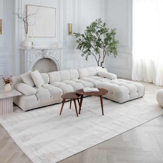 Rug Branch Astra Modern Indoor Area Rug, Cream Beige, Abstract - a large cream beige abstract area rug with subtle linear patterns, placed in a bright living room featuring a tufted cream sectional sofa, two mid-century modern wooden nesting coffee tables, a white decorative fireplace, and a tall green indoor plant near white paneled walls and sheer curtains, ideal for living room, bedroom, dining room, or kitchen.