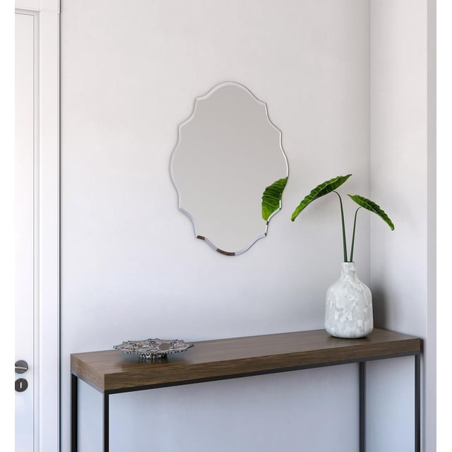 A&E Bath et Refuge de Douche Miroir Décoratif avec un Bord Biseauté Sans Cadre et une Forme Ornée, Monté sur un Mur Blanc au-Dessus d'Une table console Minimaliste avec un Dessus en Bois Foncé et des Pattes en Métal Noires, à Côté d'Un vase Texturé Blanc Tenant Trois Grandes Feuilles Vertes, Convient à la Décoration de Salle de Bain, de Chambre Ou de Couloir.