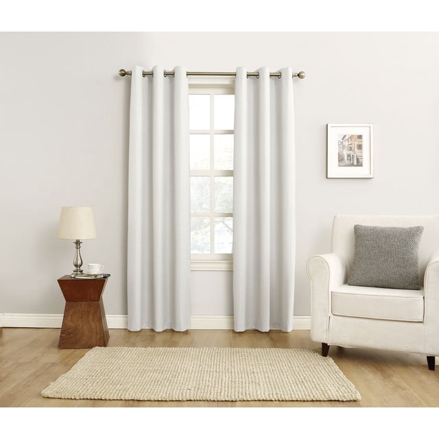 MAINSTAYS Textured Thermal-Lined Grommet Window Curtains Panel in light neutral color with woven texture and thermal lining hanging on a silver metal rod over a window in a minimalist living room with light wood flooring, beige textured rug, white armchair with gray knitted pillow, wooden side table with silver lamp and cup, and framed street art print. The thermal lining reduces energy loss and noise, ideal for enhancing room comfort.