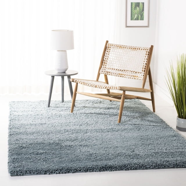 Safavieh California Solid Plush Shag Area Rug in soft grey with a dense 4.5kg/m² pile, placed under a light wood lounge chair with woven white seat and backrest, beside a small round grey side table with a white textured lamp, in a bright room with sheer curtains, a framed cactus print, and a tall green plant in a grey pot, often used to add warmth and softness to modern living spaces.