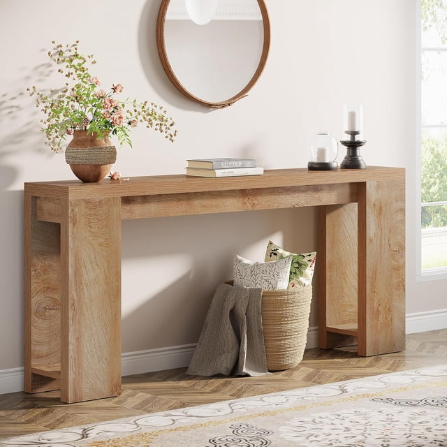 Tribesigns 70.9-inch extra long console table in light oak finish, featuring a rustic wooden tabletop and sturdy structure. The table is styled in a living room setting, placed against a light wall with a round mirror above it. Decorative items like a vase with flowers, books, and candles are displayed on the tabletop, while a woven basket with pillows and a blanket is positioned underneath. Its narrow design and versatile functionality make it suitable for use as a sofa table, entryway table, or hallway console.