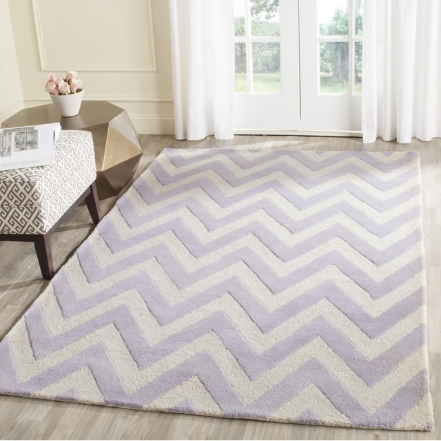 Safavieh Cambridge Kaitlyn Zig Zag Stripes Area Rug in a bright room with light wood flooring, featuring hand-tufted wool with textured high and low pile in lavender and cream zig zag stripes, styled with a patterned ottoman and geometric metallic side table, ideal for adding modern American style to home or office decor.